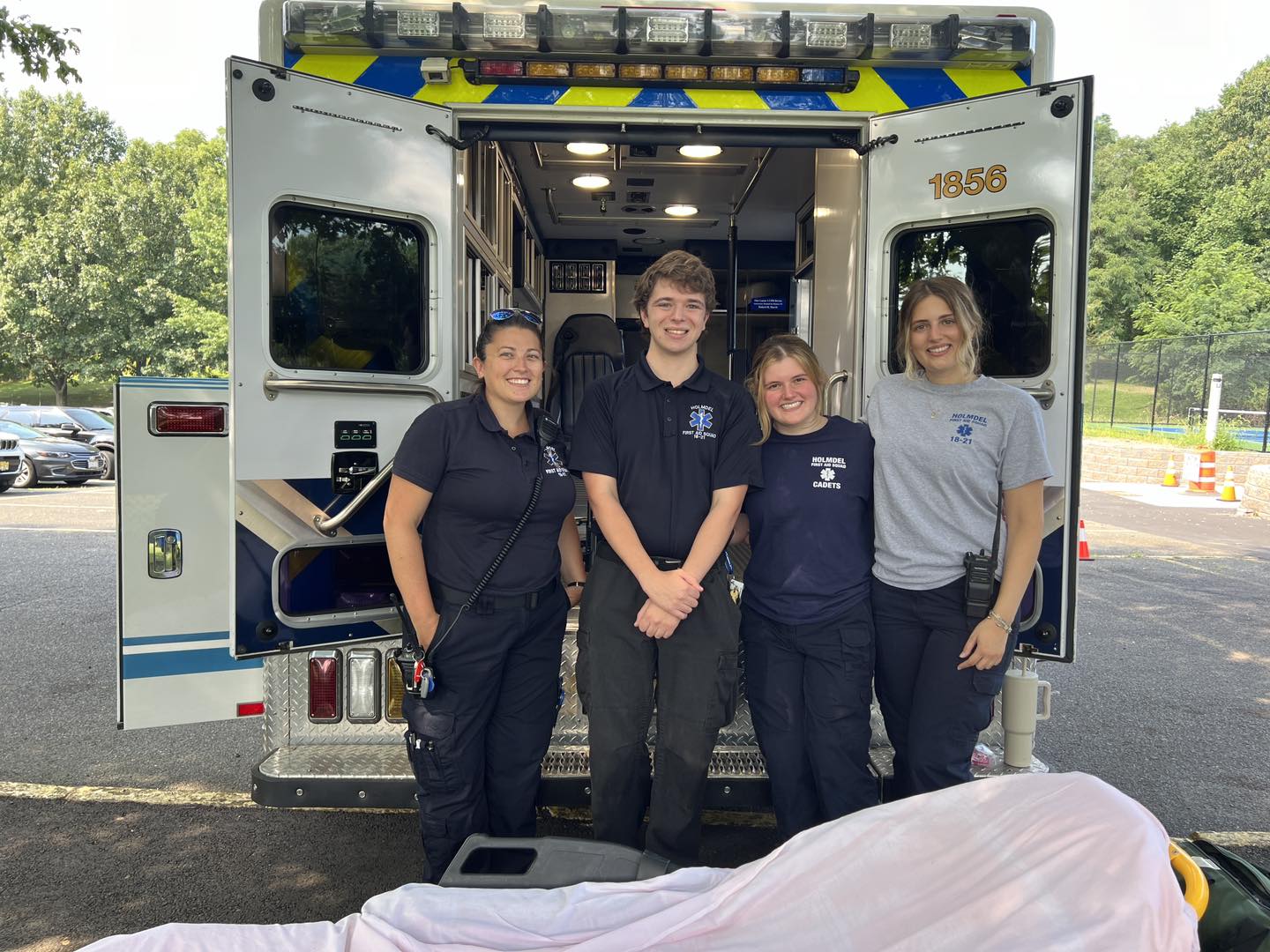 Four EMTs stand smiling in front of an open ambulance parked outdoors.