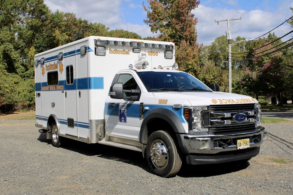 A white and blue ambulance with "Holmdel" on the side parked on a gravel surface, surrounded by trees and power lines in the background.